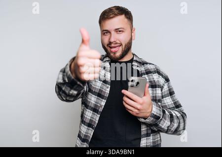 Ein Mann, der die Daumen hoch hält, während er ein Smartphone hält, lächelt. Stockfoto