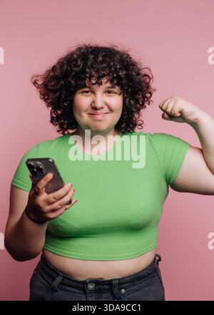 Eine fröhliche junge Frau steht zuversichtlich in einem leuchtend grünen Crop Top, beugt ihren Arm, um Kraft zu zeigen, während sie ihr Handy in der anderen Hand hält. Die Stockfoto