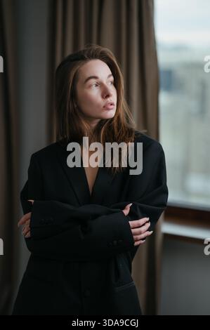 Eine junge Frau steht mit überkreuzten Armen in einem stylischen, übergroßen schwarzen Blazer. Natürliches Licht zieht durch das Fenster und hebt ihre Kontemplati hervor Stockfoto