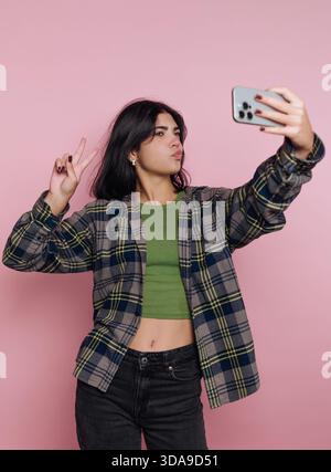 Eine junge Frau posiert für ein Selfie, macht ein Friedenszeichen und schmeckt leicht. Sie trägt ein grünes Oberteil unter einem karierten Hemd und steht vor einem hellen Stockfoto