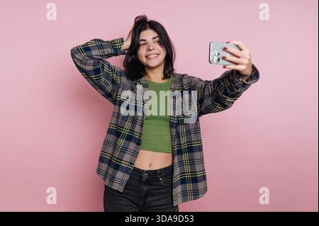 Eine junge Frau lächelt hell, während sie ein Selfie vor einer pinkfarbenen Wand macht. Sie trägt ein kariertes Hemd über einem grünen Oberteil und zeigt einen lustigen und lässigen Stall Stockfoto