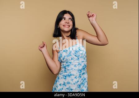 Eine junge Frau tanzt fröhlich in einem blauen Blumenkleid in einem hellen Innenraum mit warmem Hintergrund. Sie drückt Freude und Zuversicht durch h aus Stockfoto