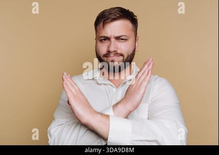 Ein Mann mit Bart und kurzen Haaren steht selbstbewusst und kreuzt seine Arme in einer Geste der Missbilligung. Er trägt ein weißes Hemd und steht gegen ein Stockfoto