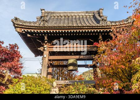 Glockenturm (Shōrō) eines buddhistischen Tempels mit einer hängenden Bronzeglocke, eingerahmt von Ahornbäumen, die im Herbst in feurigem Rot leuchten; am Fuße des Turms steht eine kleine Steinstatue, Kiso, Präfektur Nagano, Japan Stockfoto