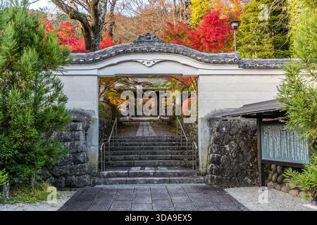 Ein weißes Tor im Stil eines Karamons mit einer Steintreppe; dahinter liegt ein Tempelgelände, das in herbstlichen Farben getaucht ist. Das Tor ist Teil eines Tempelkomplexes im Kiso-Tal in Nagano, Japan Stockfoto