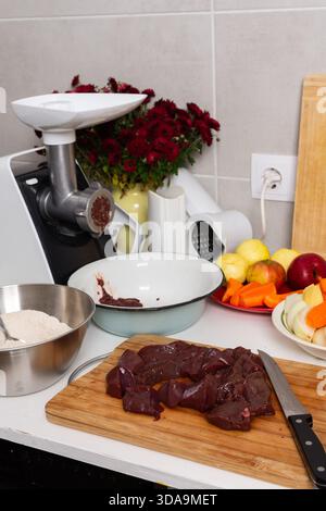 Eine Person schneidet Leber auf einem Holzbrett, während sie Leberkoteletts in einer Küche zubereitet. Frisches Gemüse und Obst stehen in der Nähe. Herbstblumen werden hineingelegt Stockfoto