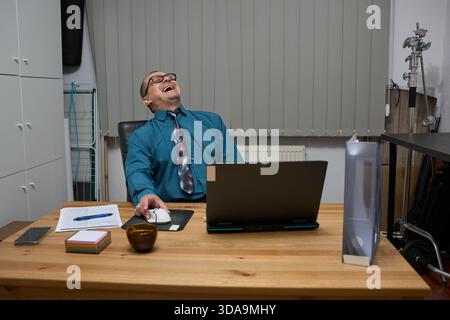 Ein glücklicher, reifer Geschäftsmann in formeller Kleidung sitzt an seinem Holztisch in seinem Heimbüro und lacht von Herzen Stockfoto