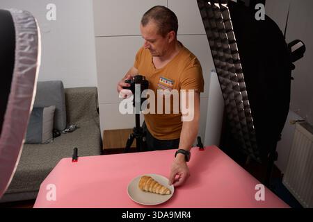 Ein fokussierter reifer Mann in einem Senf-T-Shirt steht und stellt eine Kamera auf einem Stativ ein, um einen Keks vor rosa Hintergrund zu fotografieren Stockfoto
