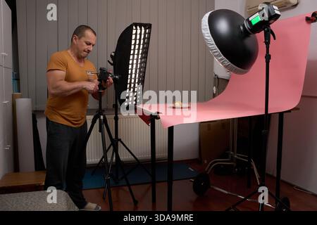 Ein fokussierter reifer Mann in einem Senf-T-Shirt steht und stellt eine Kamera auf einem Stativ ein, um einen Keks vor rosa Hintergrund zu fotografieren Stockfoto