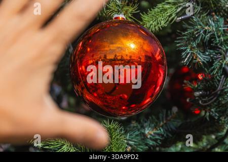 Eine Hand nähert sich einer glänzenden roten Kugel, die eine festliche Outdoor-Szene auf einem geschmückten Weihnachtsbaum reflektiert. Stockfoto