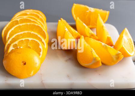 Frische Orangenscheiben und Keile auf einem Schneidebrett aus weißem Marmor. Heller Snack für Zitrusfrüchte mit saftiger Textur auf grauem Hintergrund. Stockfoto