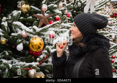 Frau, die auf einem Weihnachtsmarkt verschneite Weihnachtstanne mit Ornamenten und Lichtern sieht. Im Freien. Winter, kalte Jahreszeit. Stockfoto