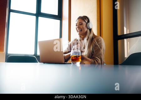 Eine junge Frau, die weiße Kopfhörer trägt, taucht mit einem Laptop in ihre Arbeit ein und sitzt in einem kreativ bunten Design am Schreibtisch. Ein Getränkeglas wird platziert Stockfoto