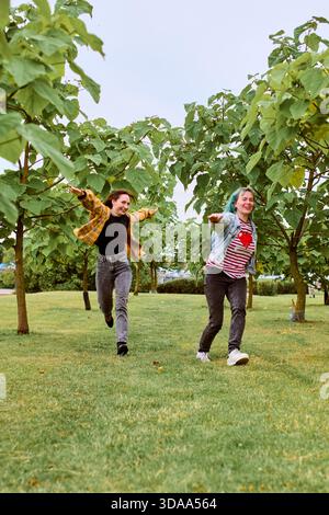 Zwei junge Frauen laufen fröhlich über einen grasbewachsenen Park mit Bäumen im Hintergrund. Beide haben ihre Arme zur Seite, lächeln und genießen Outdoor-Momente Stockfoto