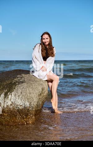 Eine junge Frau mit langen braunen Haaren und heller Haut sitzt in einem weißen Schal auf einem großen Felsen am Ostseestrand. Ruhige Wellen und helles Tageslicht sorgen für Stockfoto