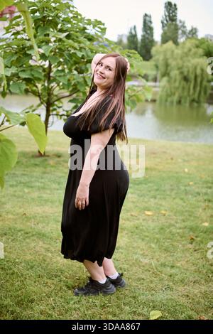 Junge Frau mit braunen Haaren in schwarzem Kleid tanzt glücklich auf dem Grasfeld. Bäume und Teich bilden einen ruhigen Hintergrund. Helles, natürliches Licht verbessert die Stockfoto