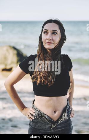 Erwachsene Frau mit langen braunen Haaren und schwarzer Spitze steht am Sandy Beach in der Nähe des Ozeans und blickt nachdenklich unter weichem Tageslicht mit ruhigem Meer i zur Seite Stockfoto