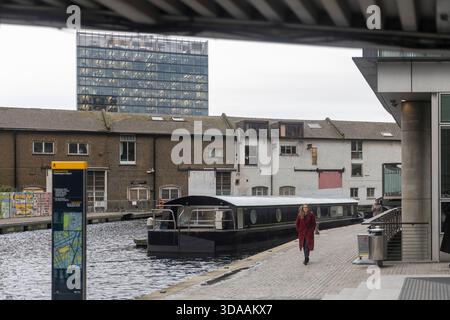 London, Großbritannien, 7. Dezember 2025, das Paddington Basin. Eine Frau in rotem Mantel, die auf einem Steinpfad neben einem Kanal entlang läuft, vorbei an einem vertäuten schwarzen Schmalz Stockfoto