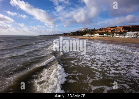 Wellen, die auf einen Sandstrand mit Gebäuden am Meer und einem bewölkten Himmel an einem Küstentag mit fernen Hügeln und Küste von Bournemouth, Großbritannien, im Dezember Stockfoto