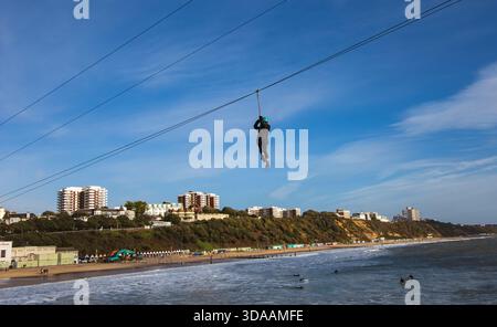 Person Seilrutsche über einem Strand mit Meereswellen und Gebäuden an der städtischen Küste unter einem klaren blauen Himmel. Bournemouth Pier Zip Dezember 2022 Stockfoto