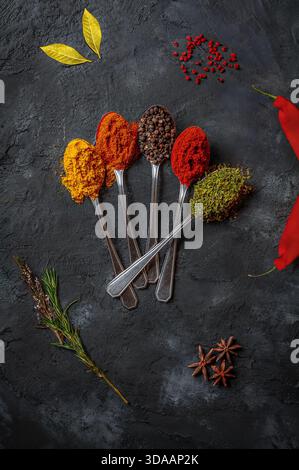 Flache Lage mit verschiedenen bunten Gewürzen, die in Löffeln auf einem dunklen, strukturierten Hintergrund angeordnet sind. Stockfoto