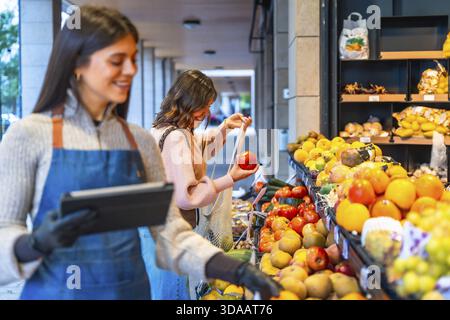 Gemüsehändler lächelt und hält ein digitales Tablet in der Hand, während eine glückliche Konsumentin frische Tomaten, Obst und Gemüse von einem Markt im Freien wählt Stockfoto