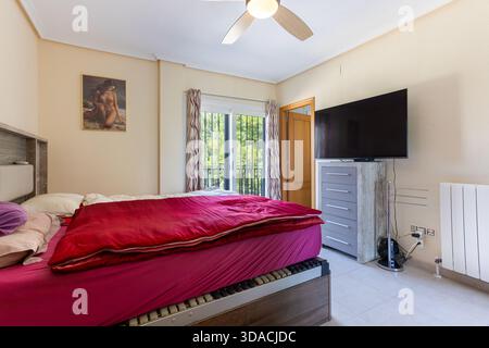 Komfortables Schlafzimmer mit roter Bettwäsche, großem Fernseher, Deckenventilator und Tageslicht. Stockfoto