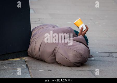 LONDON, GROSSBRITANNIEN. Februar 2025. Bettler, der auf dem Boden liegt, mit Glas in der Hand. Stockfoto