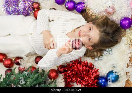 Das Kind trägt weiße Strickwaren und hält glitzernde Verzierungen, während es auf einem Pelzteppich mit Weihnachtsschmuck in der Nähe liegt Stockfoto