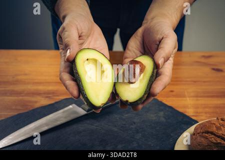 Nahaufnahme von männlichen Händen, die zwei Hälften einer Reifen frischen Avocado mit einem Samen halten. Zubereitung von gesundem Essen auf einem rustikalen Holztisch mit einem Messer und b Stockfoto