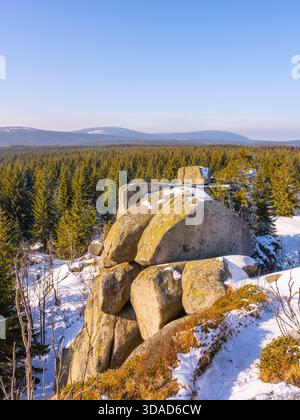 Die Sonne scheint an einem kalten Wintermorgen auf den Pytlacke-Felsen im Isergebirge. Schnee bedeckt die Landschaft, während das ruhige Dorf Jizerka in der Nähe liegt, umgeben von Bäumen. Stockfoto