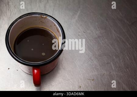 Authentischer griechischer Kaffee, serviert in einem Becher aus rotem Emaille, auf einer abgenutzten Metalloberfläche. Rustikale und minimale Komposition, ideal für redaktionelle Inhalte Stockfoto