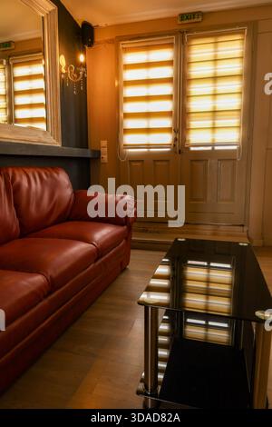 In warmen Tönen gehaltene Inneneinrichtung mit rotem Ledersofa, Glastisch und Sonnenlicht, das durch gemusterte Jalousien gefiltert wird. Stockfoto
