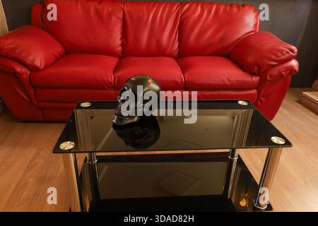 Eine moderne Wohnzimmerszene mit schwarzem Totenkopfschmuck auf einem Glastisch, im Kontrast zu einem kräftigen roten Ledersofa. Stockfoto