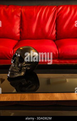 Ein elegantes schwarzes Totenkopfschmuck sitzt auf einem reflektierenden Glastisch, im Kontrast dazu steht ein lebhaftes rotes Sofa dahinter, das ein auffälliges, konzeptionelles Innenbild erzeugt. Stockfoto