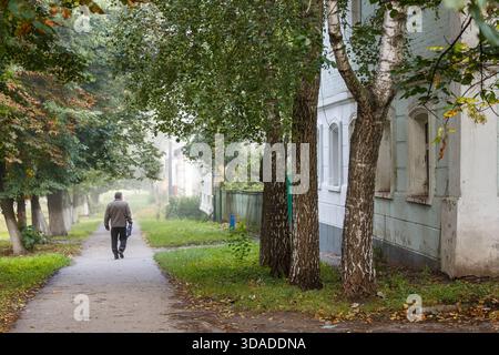 Ukraine, Romny, 09.09.2013: Ein einsamer Mann spaziert an einem ruhigen, nebeligen Tag in einem kleinen Viertel entlang eines ruhigen, baumschattigen Weges neben alten Wohnhäusern Stockfoto