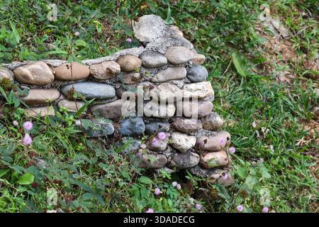 Stützmauer aus Naturstein im Garten mit rundem Flussfelsen. Diese friedliche, rustikale Kieselsteinstruktur liegt an einem grasbewachsenen Hang und ist ein heiteres Detail von lan Stockfoto