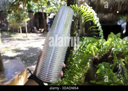 Hand hält Stapel von klarem Einweg-Plastikbecher für Party, mit grünem Outdoor-Hintergrund der Natur und des Laubs. Bereit für das Servieren kalter Getränke im an Stockfoto