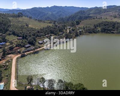Ruhiger Blick aus der Luft auf die malerische ländliche Landschaft mit grünem Wassersee, einem kleinen Dorf und einem riesigen Wald vor Berghintergrund, wunderschöner Natur Stockfoto