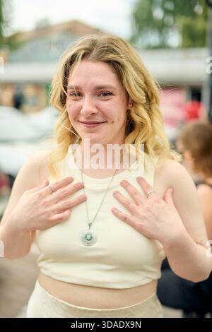 Junge blonde Frau mit lockigem Haar lächelt sanft draußen an hellem Tag. Sie trägt ein helles ärmelloses Oberteil und eine Halskette mit Anhänger und verschwommenem urb Stockfoto
