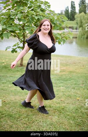 Junge Frau mit braunen Haaren in schwarzem Kleid tanzt glücklich auf dem Grasfeld. Bäume und Teich bilden einen ruhigen Hintergrund. Helles, natürliches Licht verbessert die Stockfoto