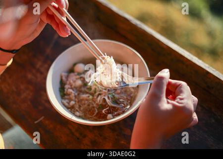 Nahaufnahme der Hand mit Essstäbchen, um eine warme Schüssel Nudelsuppe zu essen. Genießen Sie köstliche asiatische Gerichte im Freien während des sonnigen Tages auf rustikalem Holz Surfa Stockfoto