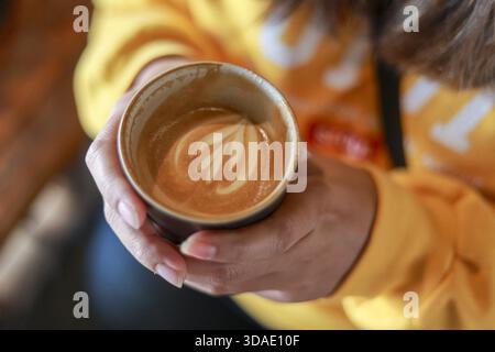 Gemütliche Frau in gelbem Pullover hält warme Tasse Kaffee mit schöner Latte Art. Entspannender Morgendrink im Café, Nahaufnahme der Hand mit Schaum und b Stockfoto