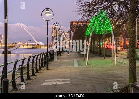 Blick in die Dämmerung entlang der Liffey Quais, Fußgänger, dekorative Straßenbeleuchtung, grüner Bogen und Beckett Bridge in der Ferne. Stockfoto