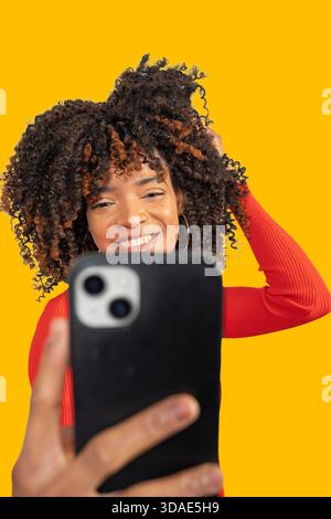 Glückliche junge Frau mit lockigem Haar, die Videos filmt oder Selfie vor gelbem Hintergrund macht Stockfoto