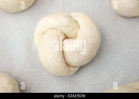 Draufsicht des Brotknotens auf Backpapier. Roher Brotteigknoten, der aufsteigt oder darauf wartet, gebacken zu werden. Traditionelles Schweizer Brotrezept namens Zopf, aber Stockfoto