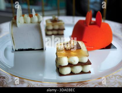 Eine detaillierte Nahaufnahme von drei Gourmet-Desserts, serviert auf einem feinen Porzellanteller. Die Auswahl umfasst einen leuchtend orangefarbenen Mousse-Kuchen mit Samt Stockfoto