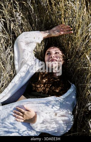 Junge Frau mit langen braunen Haaren liegt auf trockenem Roggenfeld und schirmt die Augen mit einer Hand vor Sonnenlicht ab. Natürliche Goldtöne und Schatten sorgen für eine ruhige rur Stockfoto