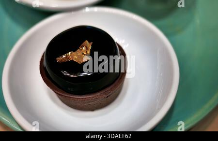 Hohe Nahaufnahme einer hochwertigen Tarte aus dunkler Schokolade mit glänzender Spiegelglasur. Das Dessert wird elegant mit einem Stück essbaren Blattgold gekrönt Stockfoto