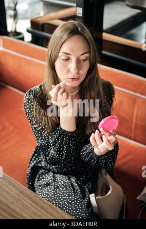Erwachsene Frau mit langen braunen Haaren sitzt auf einer rostfarbenen Bank, hält einen rosafarbenen, kompakten Spiegel und trägt Lippenstift in einer ruhigen Inneneinrichtung auf Stockfoto